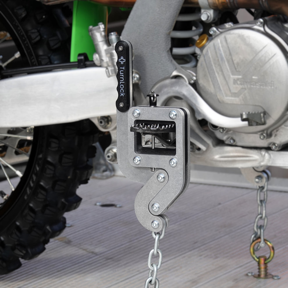 TurnLock Moto Peg Binder clamped on a dirt bike footpeg, chained to a tie-down point